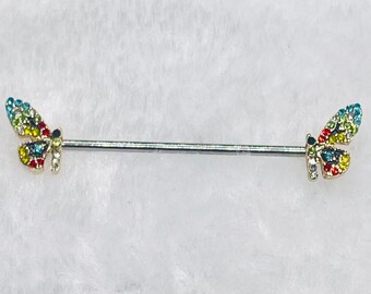 May include: A silver industrial barbell featuring butterfly charms. Each butterfly is adorned with colourful, sparkling gems in shades of blue, green, yellow, and red. The barbell is designed for body piercings.