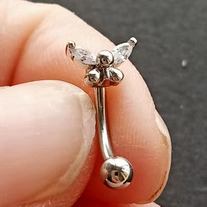 May include: A silver-colored eyebrow piercing with a curved barbell design. The top features a cluster of small, round accents and three clear, marquise-cut stones arranged in a floral pattern. The bottom has a small, shiny ball.