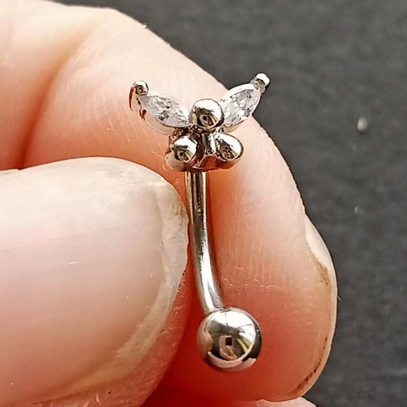 May include: A silver-colored eyebrow piercing with a curved barbell design. The top features a cluster of small, round accents and three clear, marquise-cut stones arranged in a floral pattern. The bottom has a small, shiny ball.