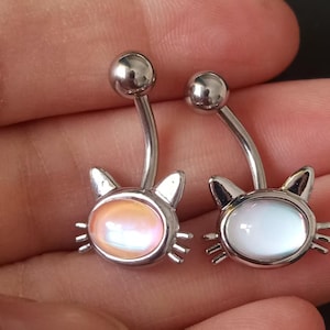 Puede incluir: Dos piercings para el ombligo plateados. Cada uno presenta un diseño de cara de gato con una barra curva y una bolita. Una cara de gato tiene una gema ovalada naranja-rosa, la otra una gema ovalada blanca.