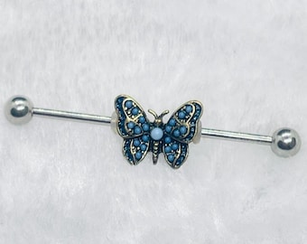 May include: An industrial barbell earring with a butterfly design. The butterfly has a gold-coloured outline and is filled with small, blue-coloured stones. The barbell is silver-coloured with a small ball on each end.