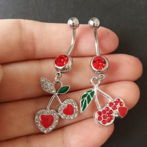 May include: Two silver belly button rings with a cherry design. Each ring features a curved barbell with a red gem at the top and a dangling cherry charm. The cherries have red gems and a green leaf.