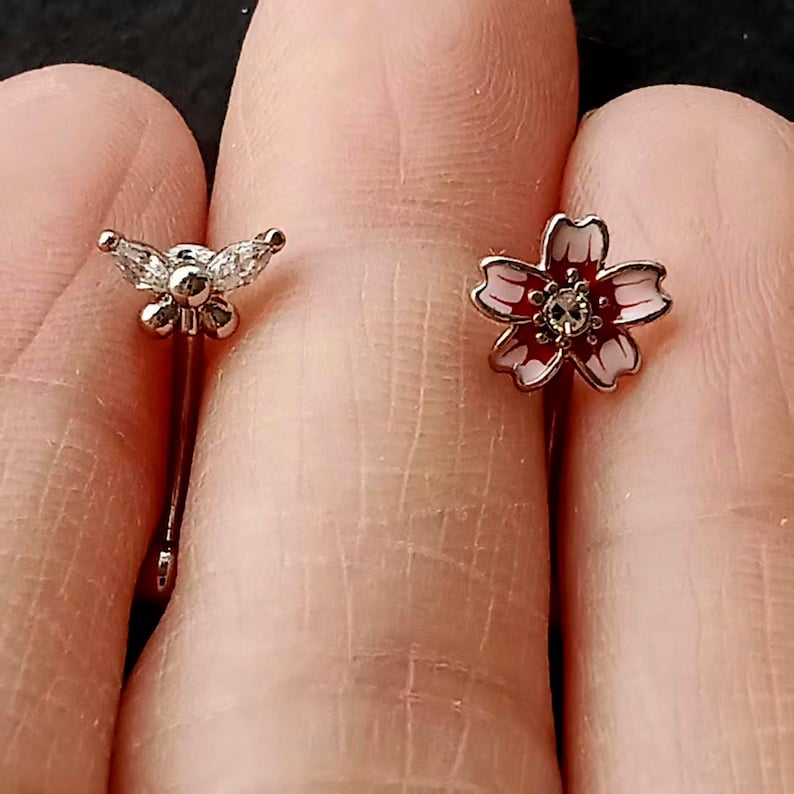 May include: Two small, decorative earrings. One earring features a butterfly design with clear, sparkling accents. The other earring has a flower design with red and white enamel petals and a central clear gem. Both earrings are silver-toned.