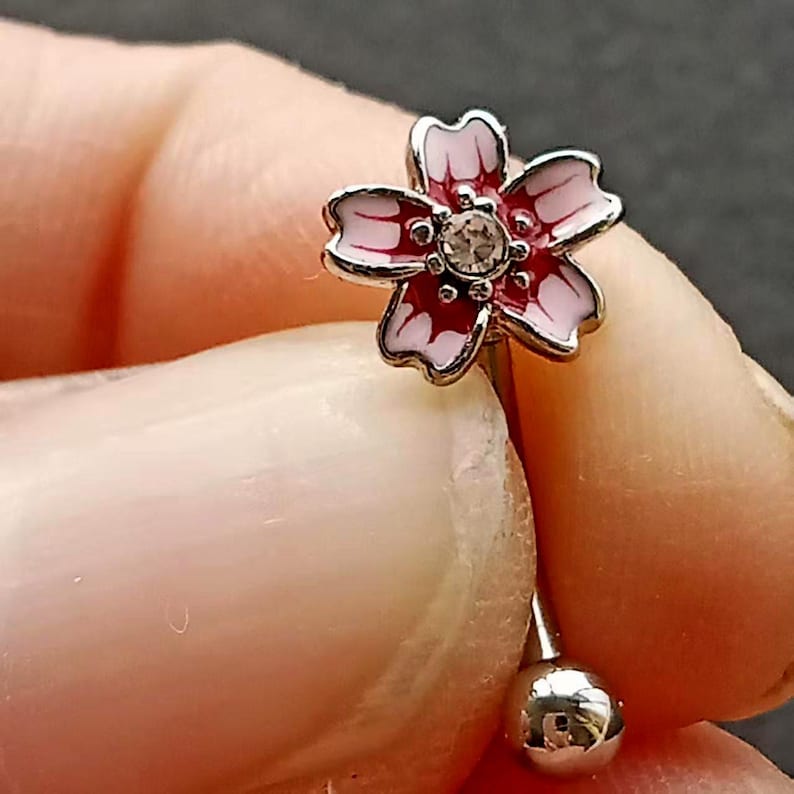 May include: A silver-colored body piercing with a pink and white flower design. The flower has a clear gem in the center and is held by a person's fingers.
