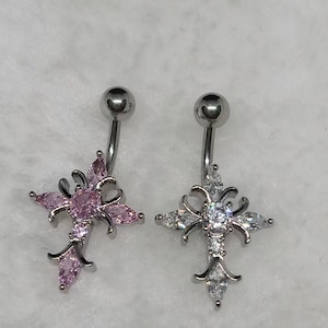 May include: Two belly button rings with cross pendants. One features pink gemstones, the other clear. Both have a silver-colored curved bar and a small ball at the top. The crosses have ornate, pointed designs.