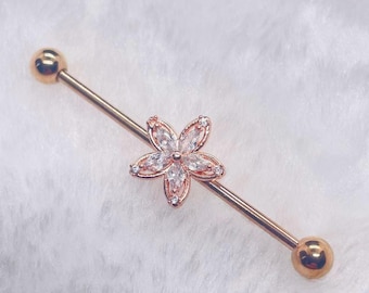 May include: Rose gold industrial barbell with a flower-shaped charm. The charm features clear crystals set in a floral design. The barbell has a smooth finish and gold-coloured ball ends.