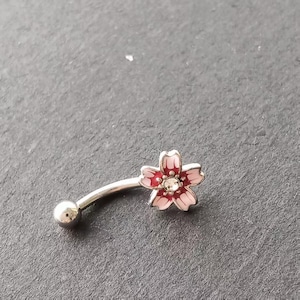 May include: A curved eyebrow ring with a flower design. The flower has pink petals with a red center and a clear gem in the middle. The metal is silver-colored, and the overall design is delicate and feminine.