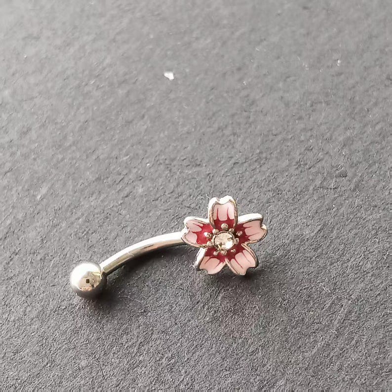 May include: A curved eyebrow ring with a flower design. The flower has pink petals with a red center and a clear gem in the middle. The metal is silver-colored, and the overall design is delicate and feminine.
