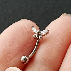 May include: A curved eyebrow piercing with a flower design. The jewelry is silver-toned, with a small ball on one end and a flower with three clear, teardrop-shaped gems on the other. The flower has a cluster of small, round petals.