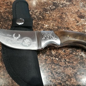 Könnte beinhalten: Ein Jagdmesser mit gebogener Klinge und Holzgriff, das auf einer schwarzen Scheide liegt. Die Klinge zeigt eine gravierte Hirschszene und die Worte "AMERICAN HUNTING". Der Griff ist dunkelbraun.