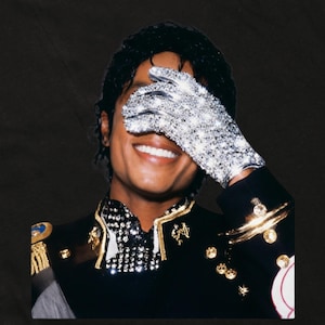 Puede incluir: Un retrato de una persona sonriente con una chaqueta negra con ribetes dorados y un guante plateado brillante que cubre sus ojos. La imagen tiene un fondo oscuro.