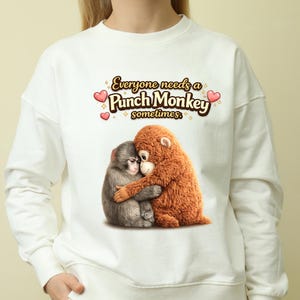 May include: White sweatshirt featuring a graphic of two hugging monkeys. One monkey is gray, the other is brown. The text "Everyone needs a Punch Monkey sometimes" is above the monkeys in a playful font with heart and star accents.