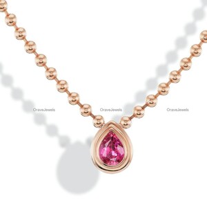May include: A rose gold necklace featuring a delicate ball chain and a single pear-shaped pink gemstone pendant set in a smooth rose gold bezel. The design includes the text OraveJewels subtly repeated along the chain links in this jewelry piece.