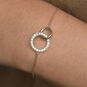 May include: A gold chain bracelet featuring two interlocking circular charms. One circle is plain polished gold, while the other is slightly larger and encrusted with a series of small, clear, sparkling stones set along its entire outer circumference.