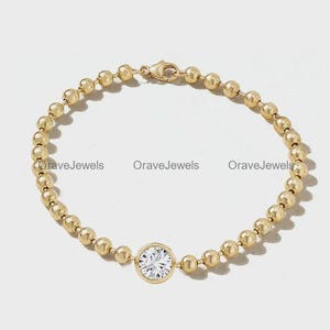 May include: A gold bead chain bracelet featuring a single round, clear, faceted stone set in a gold bezel. The bracelet includes a lobster claw clasp and is marked with the text OraveJewels repeated three times across the centre of the image.