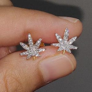 May include: A pair of stud earrings shaped like cannabis leaves, featuring a metallic gold-tone base and a surface covered in small, clear, sparkling stones that catch the light, creating a bright and reflective appearance on each leaf.