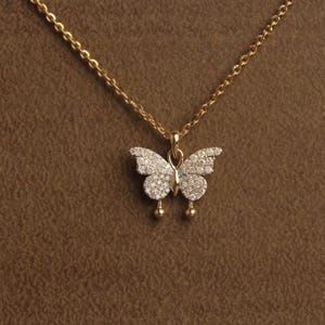 Può includere: Una collana a catena dorata con un ciondolo a forma di farfalla. Le ali della farfalla sono incastonate con numerose piccole pietre chiare e scintillanti. Il ciondolo include due piccole sfere dorate appese alle ali inferiori.