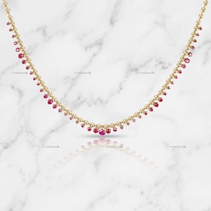 Puede incluir: Un collar de cadena dorada que presenta una serie de pequeñas esferas de oro pulido intercaladas con diminutos rubíes de color rojo vibrante. El collar está dispuesto en un diseño curvo en forma de U, con los rubíes colgando de las cuentas.