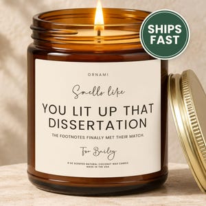 May include: A lit candle in an amber glass jar with a gold lid. The label reads: ORNAMI, Smells like YOU LIT UP THAT DISSERTATION, THE FOOTNOTES FINALLY MET THEIR MATCH., For Bailey, 9 OZ SCENTED NATURAL COCONUT WAX CANDLE, MADE IN THE USA.