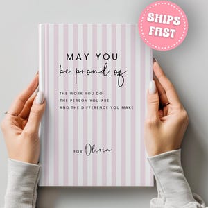 May include: A notebook with a pink and white striped cover. The cover features the text "MAY YOU be proud of THE WORK YOU DO THE PERSON YOU ARE AND THE DIFFERENCE YOU MAKE FOR Olivia."