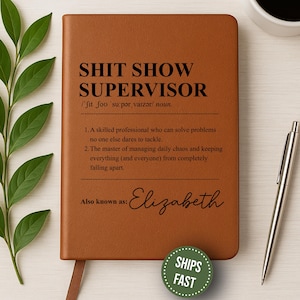 May include: A brown leather-bound notebook with the text "SHIT SHOW SUPERVISOR" in bold black letters. The notebook includes a definition and the name "Elizabeth" in cursive. A silver pen and a coffee cup are also visible.