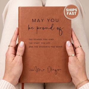 May include: A rectangular, tan-colored notebook with a textured cover. The front features black text that reads: MAY YOU be proud of THE SCHOOL YOU LEAD THE STAFF YOU LIFT AND THE STUDENTS YOU SERVE FOR Mr. Okafor. A circular sticker says SHIPS FAST.
