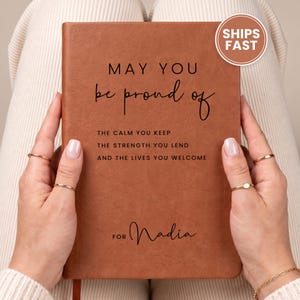 May include: A rectangular brown notebook with a textured cover featuring black text that reads: MAY YOU be proud of THE CALM YOU KEEP THE STRENGTH YOU LEND AND THE LIVES YOU WELCOME FOR Nadia. The notebook has a smooth, matte finish and a simple design.
