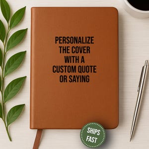 May include: A brown leather journal with the text "PERSONALIZE THE COVER WITH A CUSTOM QUOTE OR SAYING" in black. A silver pen rests on the right. A green sticker with the words "SHIPS FAST" is at the bottom. Green leaves are on the left.