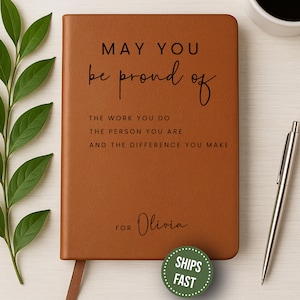 May include: A brown leather journal with the text "MAY YOU be proud of THE WORK YOU DO THE PERSON YOU ARE AND THE DIFFERENCE YOU MAKE" and "FOR Olivia". A silver pen and a green "SHIPS FAST" sticker are also visible.