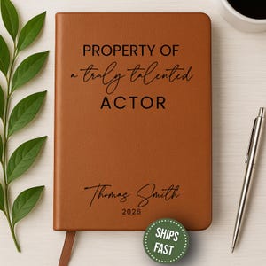 May include: A brown leather-bound journal with the text "PROPERTY OF a truly talented ACTOR" and the name "Thomas Smith" with the year "2026" printed on the cover. A silver pen and a cup of coffee are on the right. Green leaves are on the left.