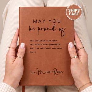 May include: A rectangular brown notebook with a textured cover featuring black printed text that reads: MAY YOU be proud of THE CHILDREN YOU FEED THE NAMES YOU REMEMBER AND THE WELCOME YOU GIVE DAILY FOR Miss Rosa.