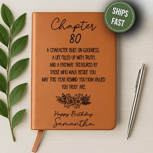 May include: A tan leather journal with the text "Chapter 80" and a personalized birthday message. The journal includes a ribbon bookmark and is accompanied by a silver pen. A green circular sticker with the words "SHIPS FAST" is in the upper right corner.