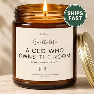 May include: A lit candle in an amber glass jar with a gold lid. The label reads ORNAMI, Smells like A CEO WHO OWNS THE ROOM, SPARKLE WITH AUTHORITY, For Hana, and 9 OZ SCENTED NATURAL COCONUT WAX CANDLE MADE IN THE USA.