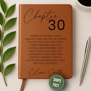 May include: A brown leather journal with the words "Chapter 30" and inspirational text. The journal includes a ribbon bookmark and the name "Olivia Smith". A silver pen and a cup of coffee are next to the journal.