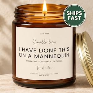 May include: A lit scented candle in an amber glass jar with a gold lid. The white label features the text: ORNAMI, Smells like I HAVE DONE THIS ON A MANNEQUIN, SIMULATION CONFIDENCE UNLOCKED, For Amelia, 9 OZ SCENTED NATURAL COCONUT WAX CANDLE.