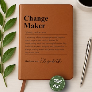 May include: A brown leather journal with the words "Change Maker" and a definition printed on the cover. The journal also includes the name "Elizabeth" and a "Ships Fast" sticker. A silver pen and a cup of coffee are next to the journal.
