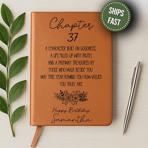 May include: A brown leather journal with the text "Chapter 37" and a personalized birthday message. The journal includes a floral design and a ribbon bookmark. A silver pen rests beside the journal.