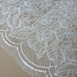 Może przedstawiać: Zbliżenie białej koronkowej tkaniny kwiatowej z falistym brzegiem. Koronka ma szczegółowe wzory liści i kwiatów, tworząc delikatny i skomplikowany wzór. Tkanina jest umieszczona na neutralnym beżowym tle.