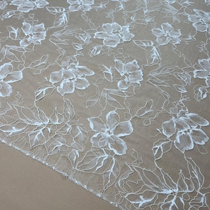 Op de afbeelding: Witte bloemenkanten stof met een delicaat, opengewerkt ontwerp. Het kant heeft een herhalend patroon van gedetailleerde bloemen en bladeren, waardoor een textuur ontstaat. De stof wordt waarschijnlijk gebruikt voor bruidskleding of andere decoratieve doeleinden.