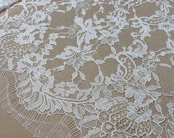 May include: Close-up of white floral lace fabric. The lace features a detailed pattern of flowers and leaves, with a scalloped edge. The fabric is laid on a tan surface, showcasing the intricate design and texture of the lace.