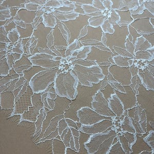 Op de afbeelding: Close-up van witte bloemenkanten stof. Het delicate kant heeft een herhalend patroon van grote, open bloemen met gedetailleerde bloemblaadjes en bladeren. Het kant staat tegen een neutrale beige achtergrond, wat het ingewikkelde ontwerp benadrukt.