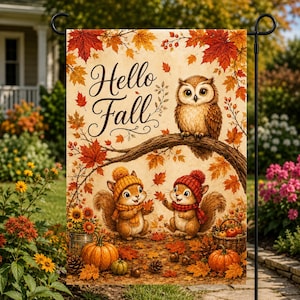 Op de afbeelding: Een rechthoekige tuin vlag met de tekst Hello Fall in zwarte schrijfletters. Het ontwerp toont een bruine uil op een boomtak boven twee eekhoorns met gebreide mutsen en sjaals, omringd door oranje esdoornbladeren en pompoenen.