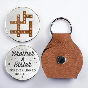 May include: A set of personalized keychains and accessories. One keychain features a brown leather holder with a silver ring. Two silver-colored round pendants have engraved text and designs. One pendant has a Scrabble-style design with names.