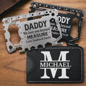 Può includere: Tre carte multi-utensili e un portafoglio in pelle nera. Una carta recita "DADDY We love you beyond MEASURE Love, Sophia & David." Gli strumenti includono righelli, apribottiglie e taglierini. Il portafoglio è impresso con il nome "MICHAEL".