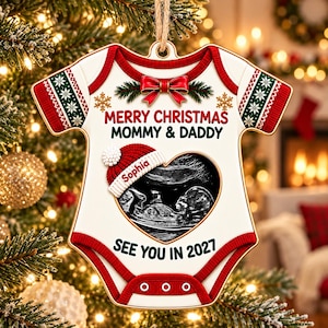May include: A white baby onesie-shaped Christmas ornament featuring a red bow, green and red patterned sleeves, and a heart-shaped ultrasound image. Text reads: MERRY CHRISTMAS MOMMY & DADDY, Sophia, and SEE YOU IN 2027, with a red and white knit hat.
