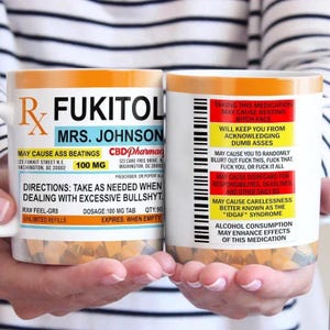 Op de afbeelding: Twee witte keramische mokken met humoristische receptlabelontwerpen. De mokken bevatten vette tekst, waaronder "FUKITOL" en "MRS. JOHNSON", samen met waarschuwingen en instructies. Het ontwerp bootst een medicijnfles na, met een speelse en oneerbiedige toon.