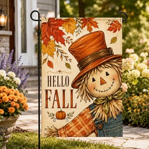 Puede incluir: Una bandera de jardín rectangular con un espantapájaros que lleva un sombrero de copa naranja y una camisa a cuadros. El texto HELLO FALL está impreso en letras marrones. El diseño incluye una pequeña calabaza naranja y hojas de arce.