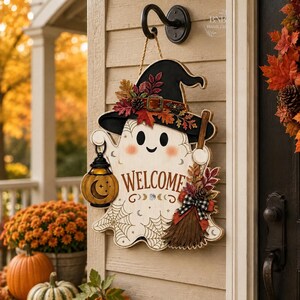 May include: A wooden ghost-shaped door sign featuring a black witch hat, a pumpkin lantern, and a broomstick. The ghost has a white body with spiderweb patterns, pink cheeks, and the word WELCOME printed in brown letters with moon phase icons.