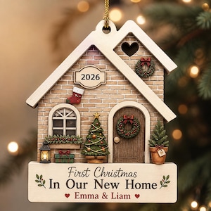 May include: A house-shaped Christmas ornament featuring a brick facade, a 2026 plaque, a stocking, a tree, and a wreath. The base displays the text First Christmas In Our New Home, Emma & Liam, with small heart icons and green leaf accents.