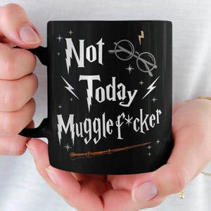 以下が含まれることがあります： 黒いセラミックマグカップ。白い文字で「Not Today Muggle f*cker」と書かれており、稲妻、メガネ、魔法の杖のイラストが描かれています。 マグカップは人が持っています。
